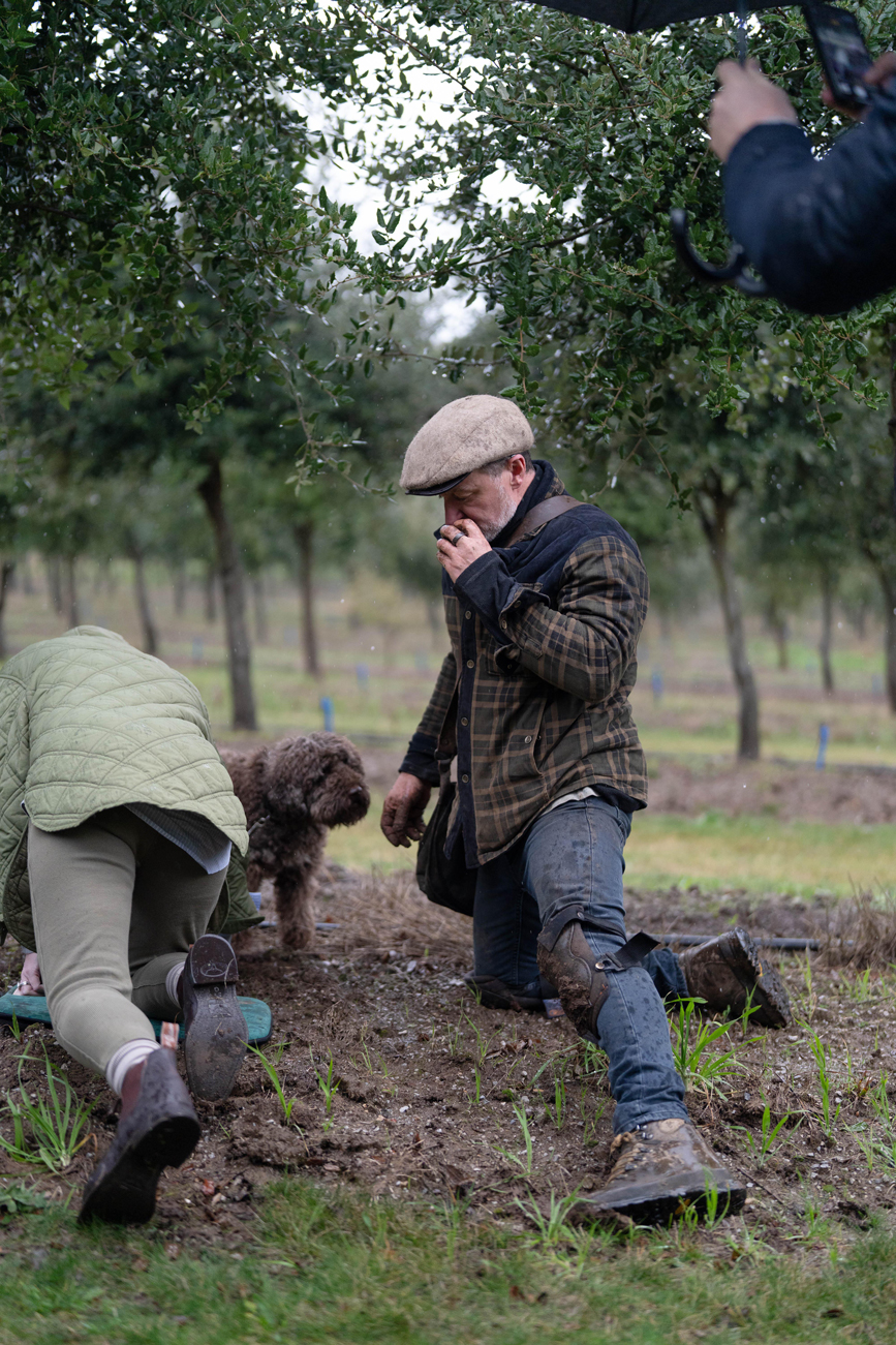 Two people and a dog kneeling and leaning forward in a muddy orchard with trees in the background.