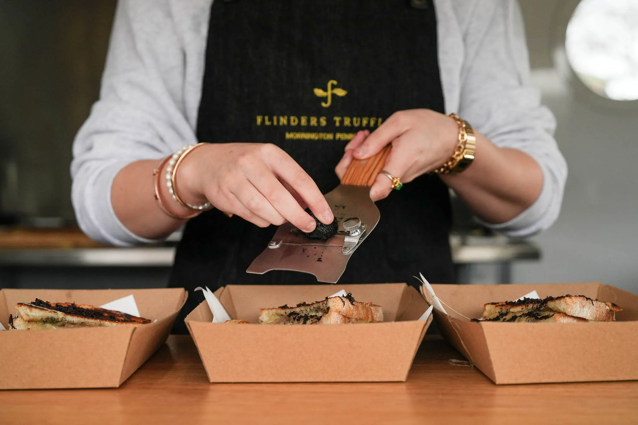 Person wearing Flinders Truffles apron grating black truffle over sandwiches in cardboard trays. Flinders Truffles Toasties