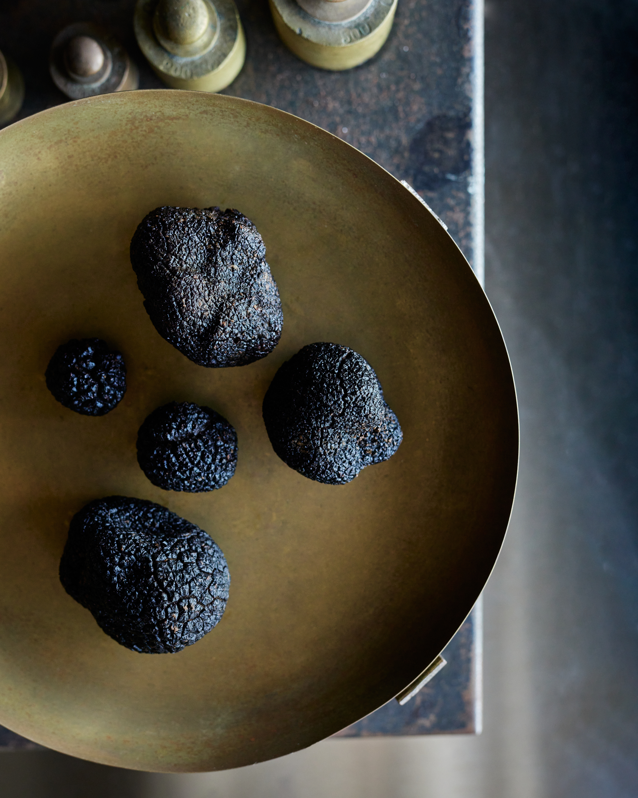 Five black truffles of varying sizes on a round brass plate on a rustic table.