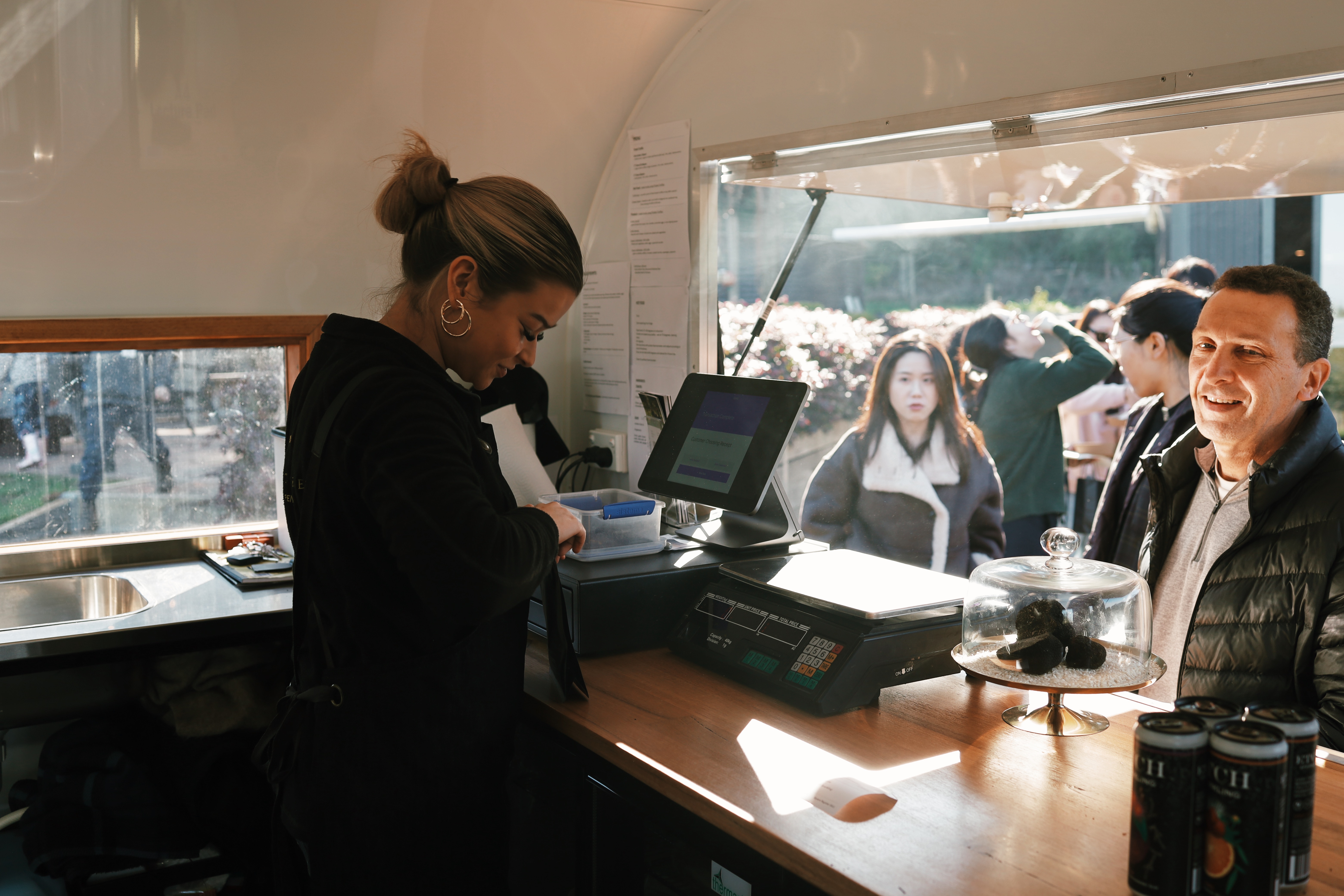 Woman purchasing at a Flinders Truffles Van with truffles under a glass dome and several beverage cans nearby.