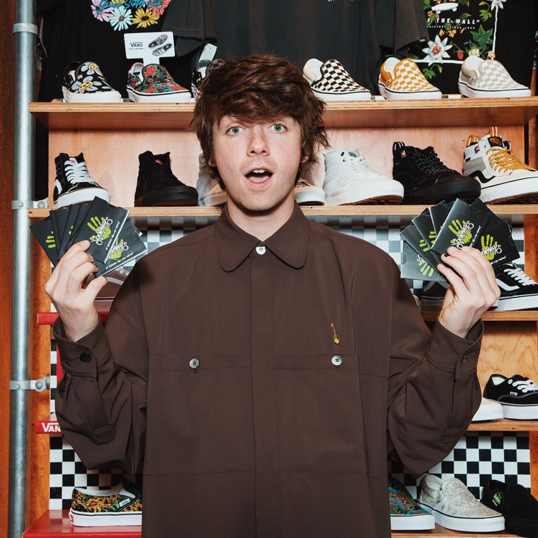 Young man with brown hair in a brown shirt holding multiple black cards with green handprints, standing in front of shelves displaying Vans shoes and apparel.