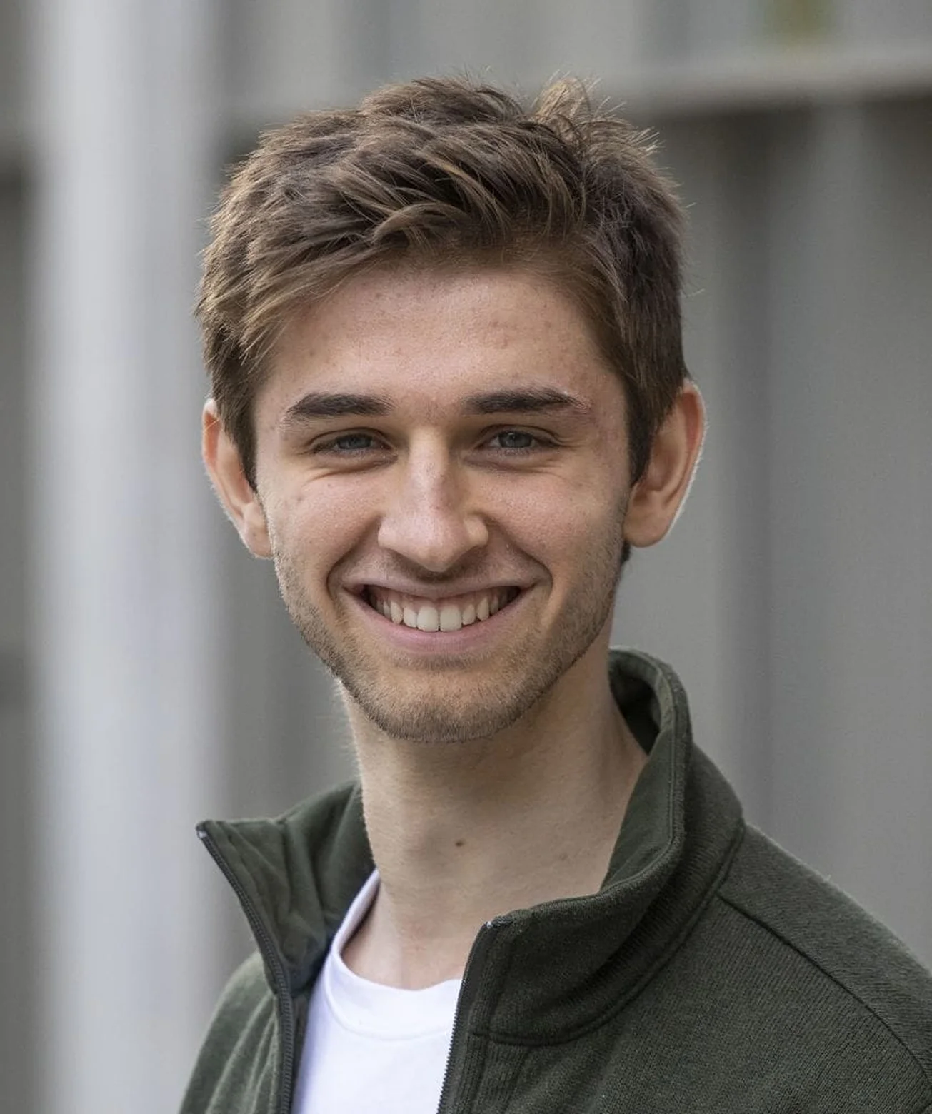 Junger Mann mit braunem Haar und grünem Pullover lächelt in die Kamera.