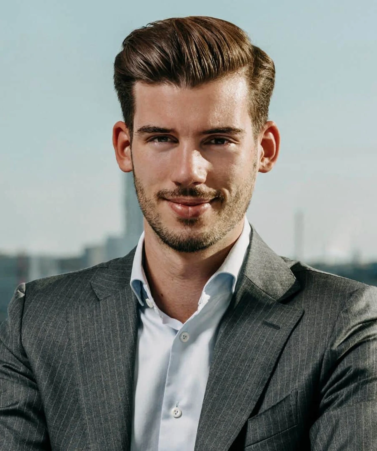 Ein lächelnder junger Mann in einem grauen Anzug und weißem Hemd vor einem städtischen Hintergrund.