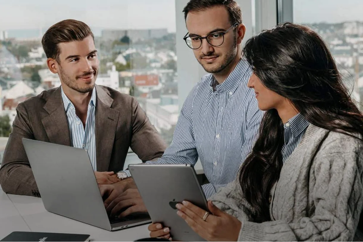 Drei Personen in einem Büro besprechen sich, zwei mit Laptop und eine mit Tablet.