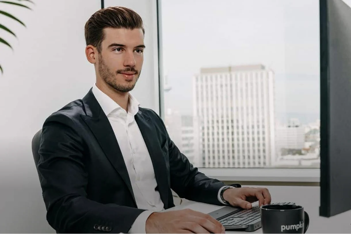 Mann im Anzug sitzt am Schreibtisch und arbeitet an einem Computer in einem Büro mit Fenster.