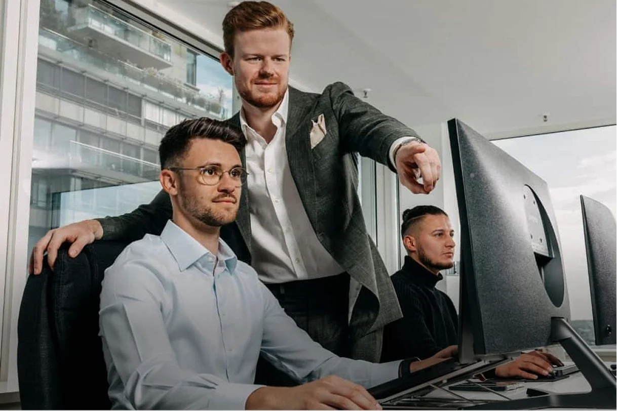 Ein Mann zeigt einem anderen am Computerbildschirm etwas, während ein dritter Mann neben ihnen arbeitet.