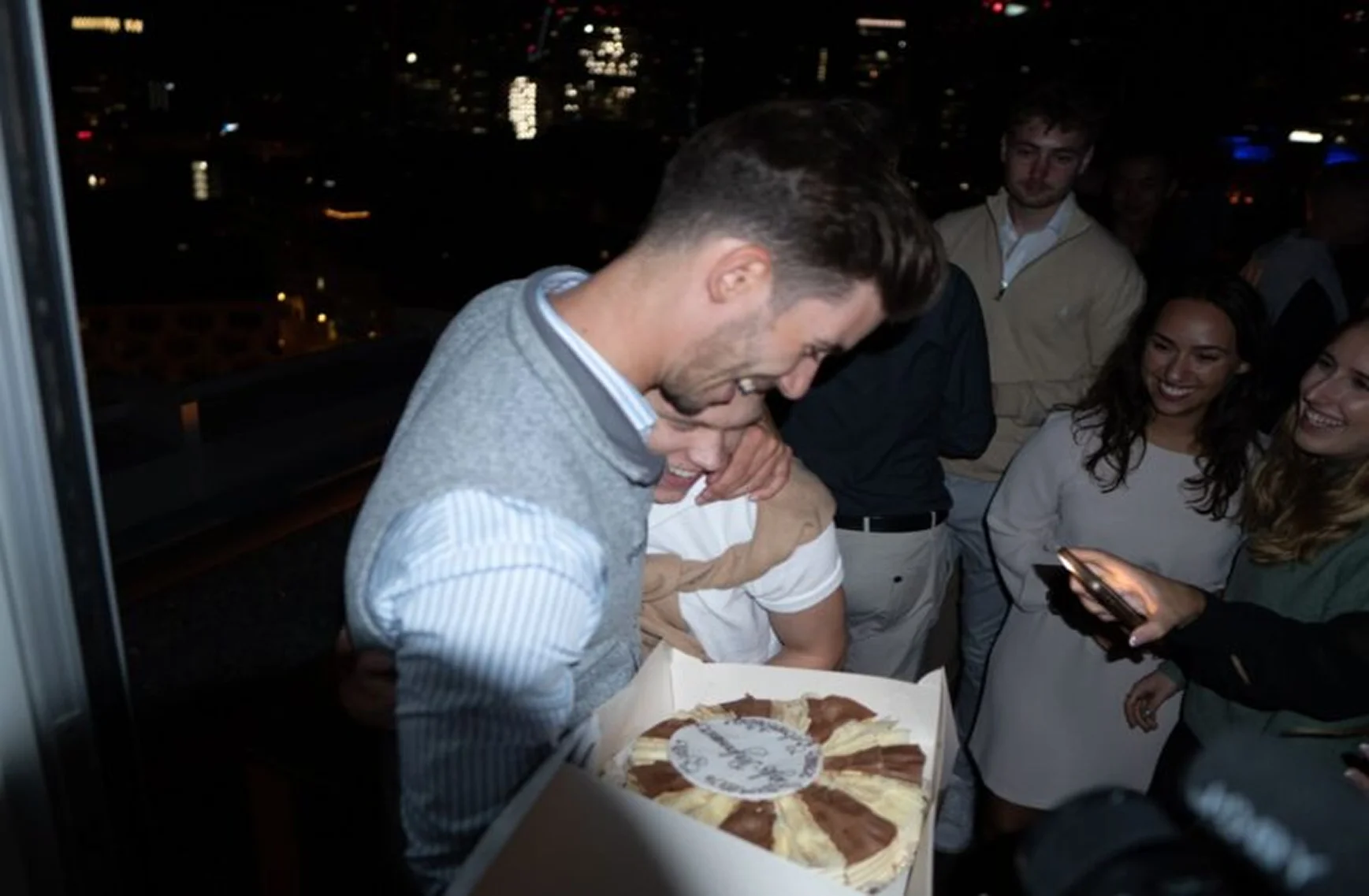 Menschen feiern nachts draußen, ein Mann hält einen Kuchen, alle lachen und schauen zum Smartphone.