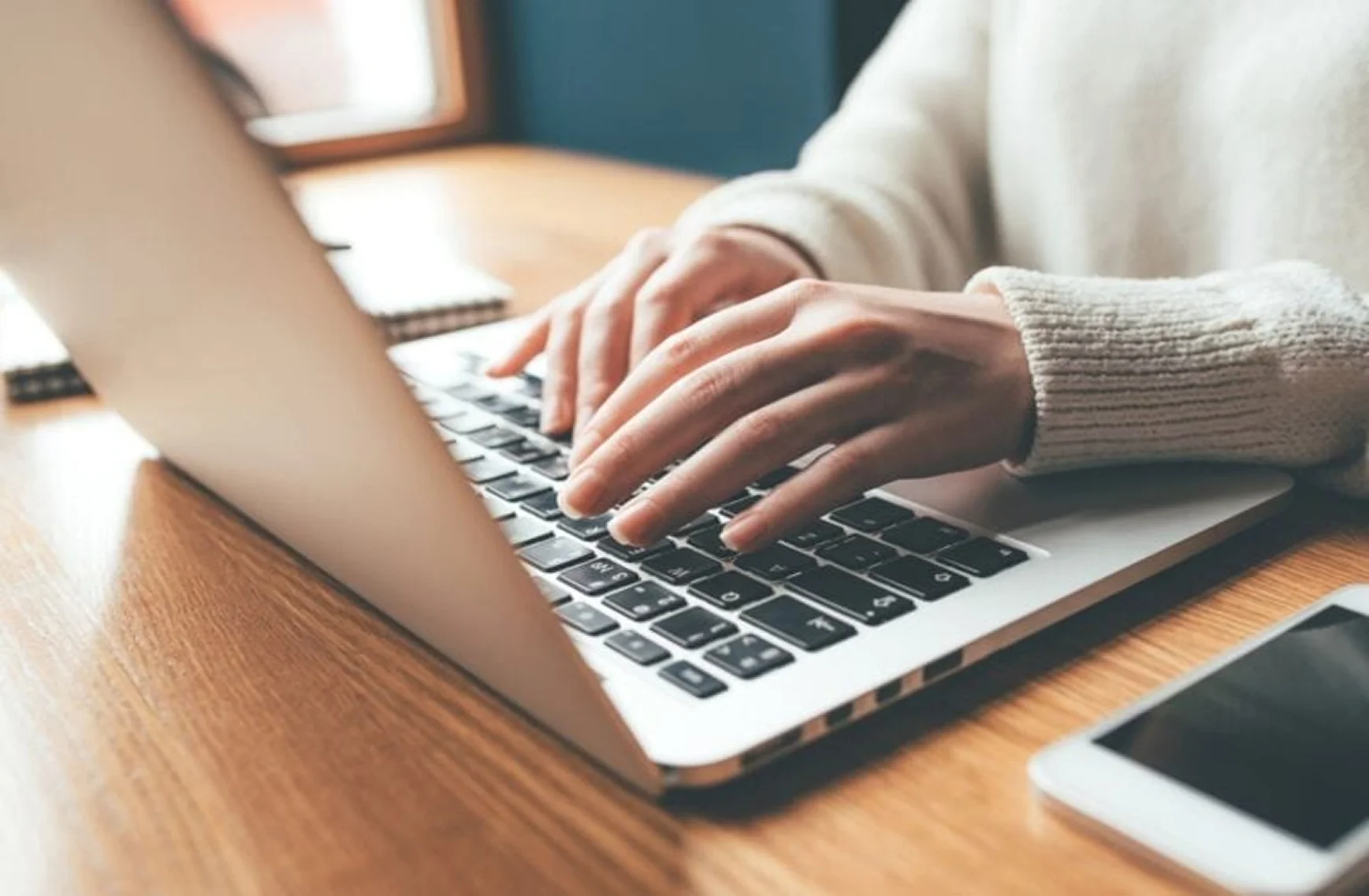 Person tippt auf Laptop-Tastatur auf einem Holztisch mit Smartphone daneben.