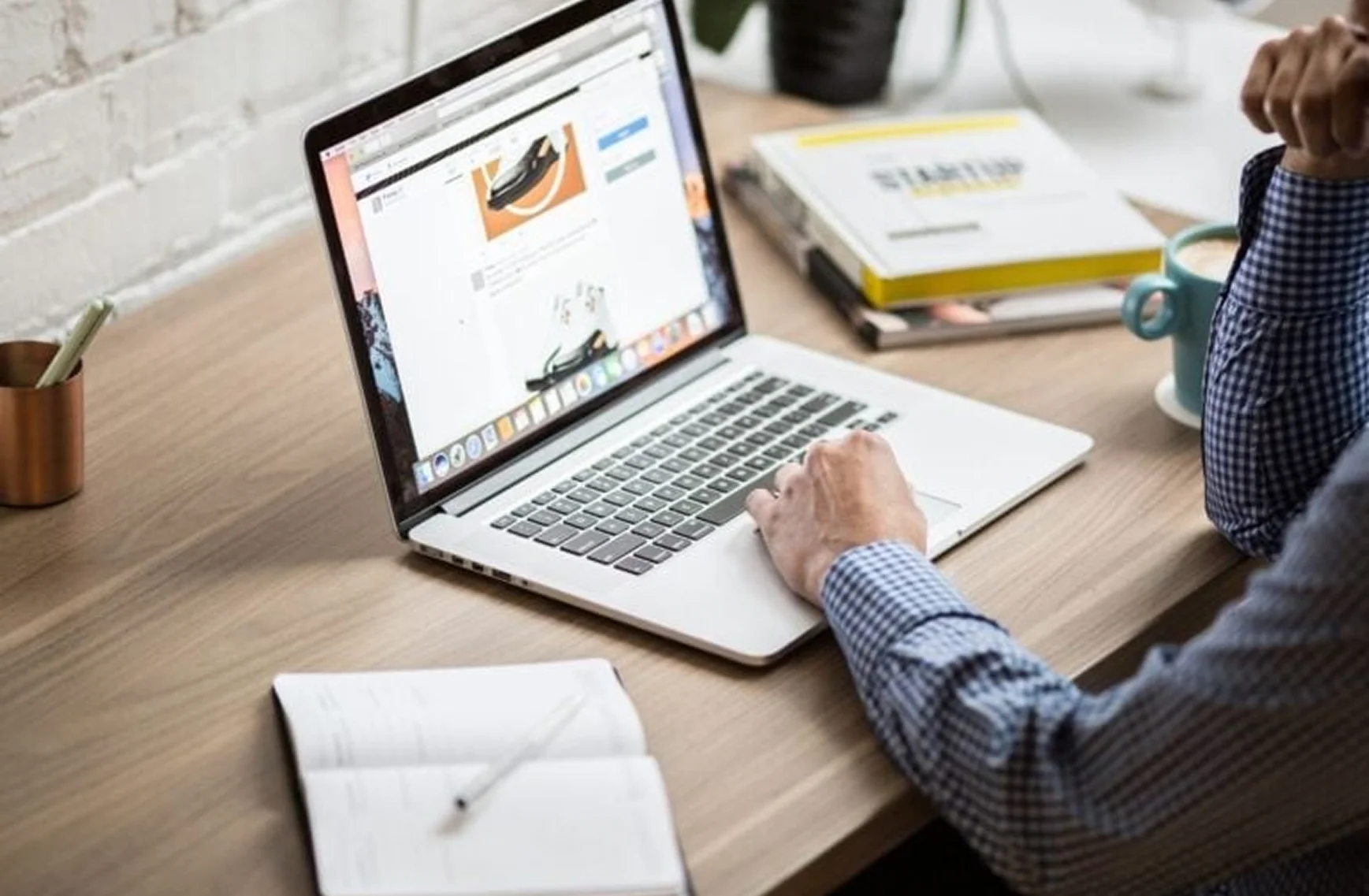 Person arbeitet an einem Laptop an einem Schreibtisch mit offenem Notizbuch und Kaffeetasse.