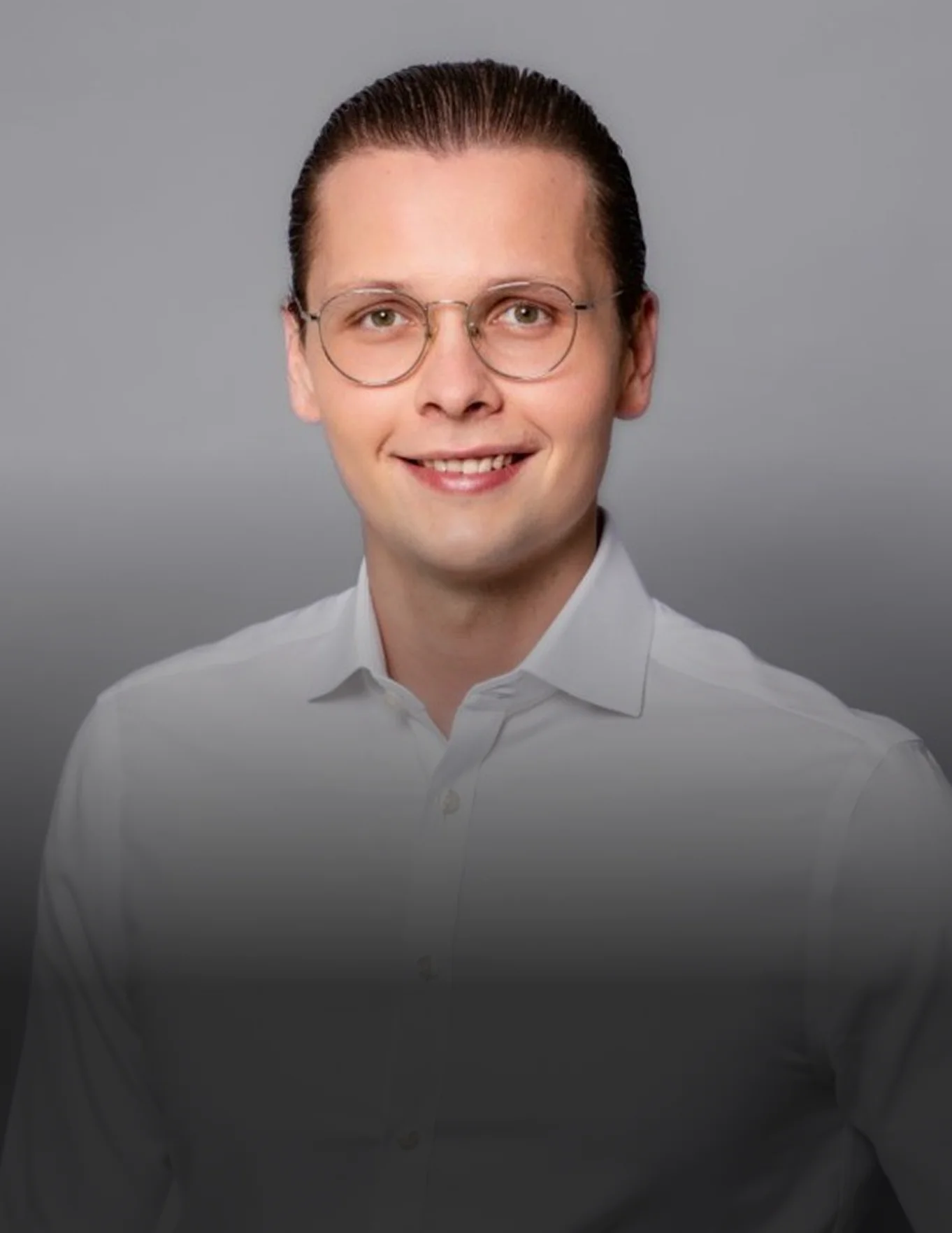 Ein lächelnder junger Mann mit Brille und zurückgekämmtem Haar trägt ein weißes Hemd vor grauem Hintergrund.