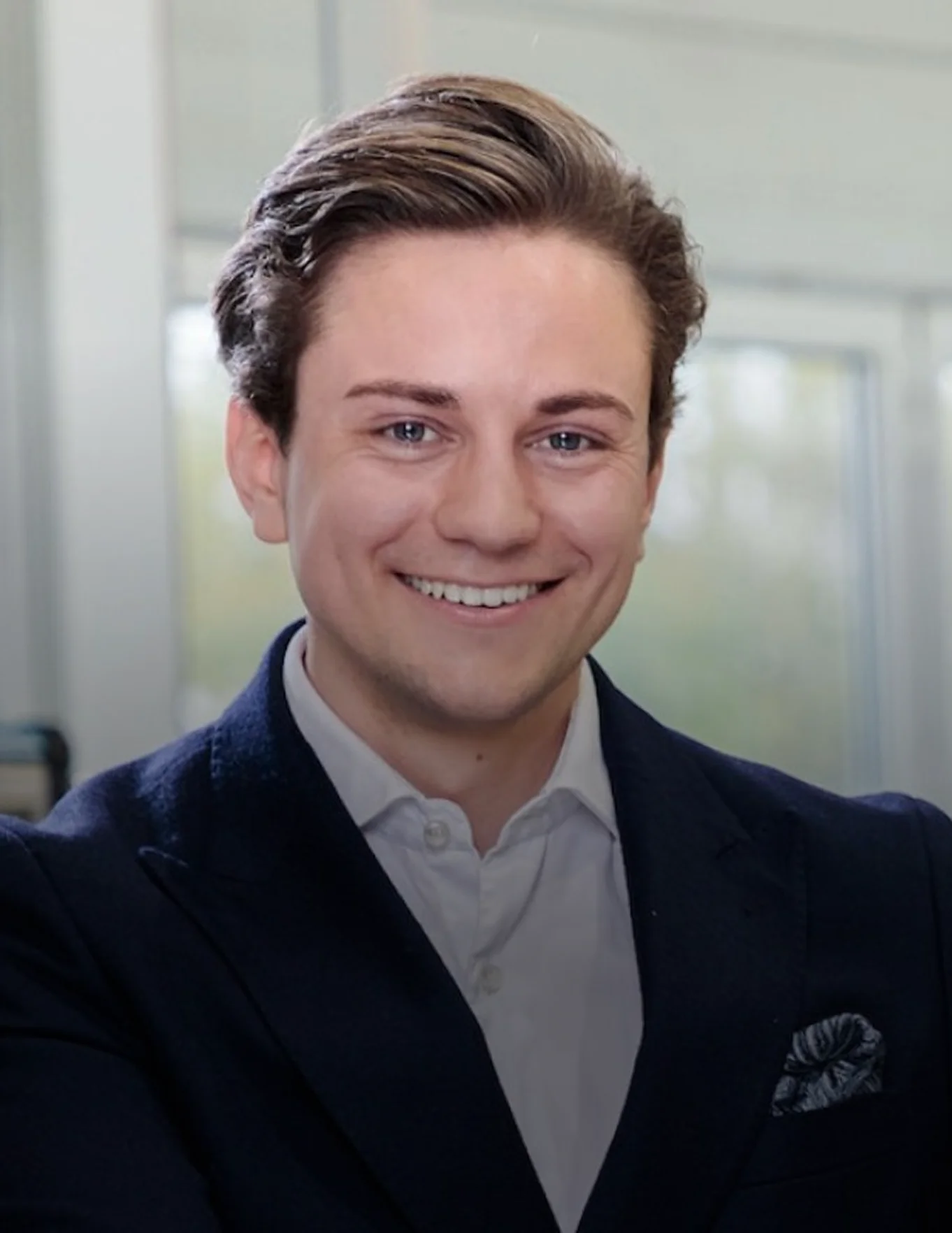 Junger Mann in dunkelblauem Anzug und weißem Hemd lächelt in einem hellen Büro.
