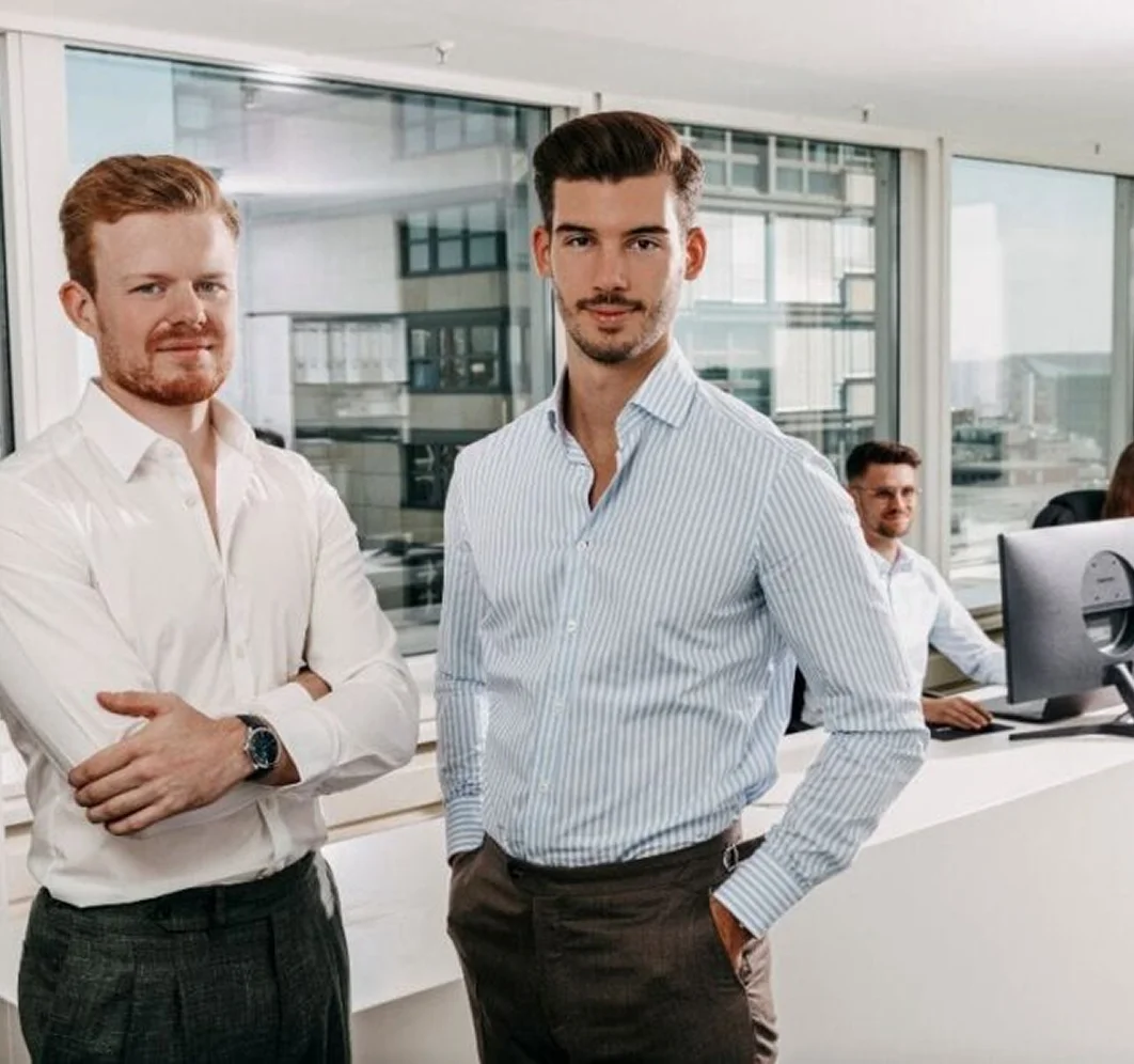 Zwei Männer stehen im Büro, einer mit weißem Hemd, der andere mit blau-weiß gestreiftem Hemd.