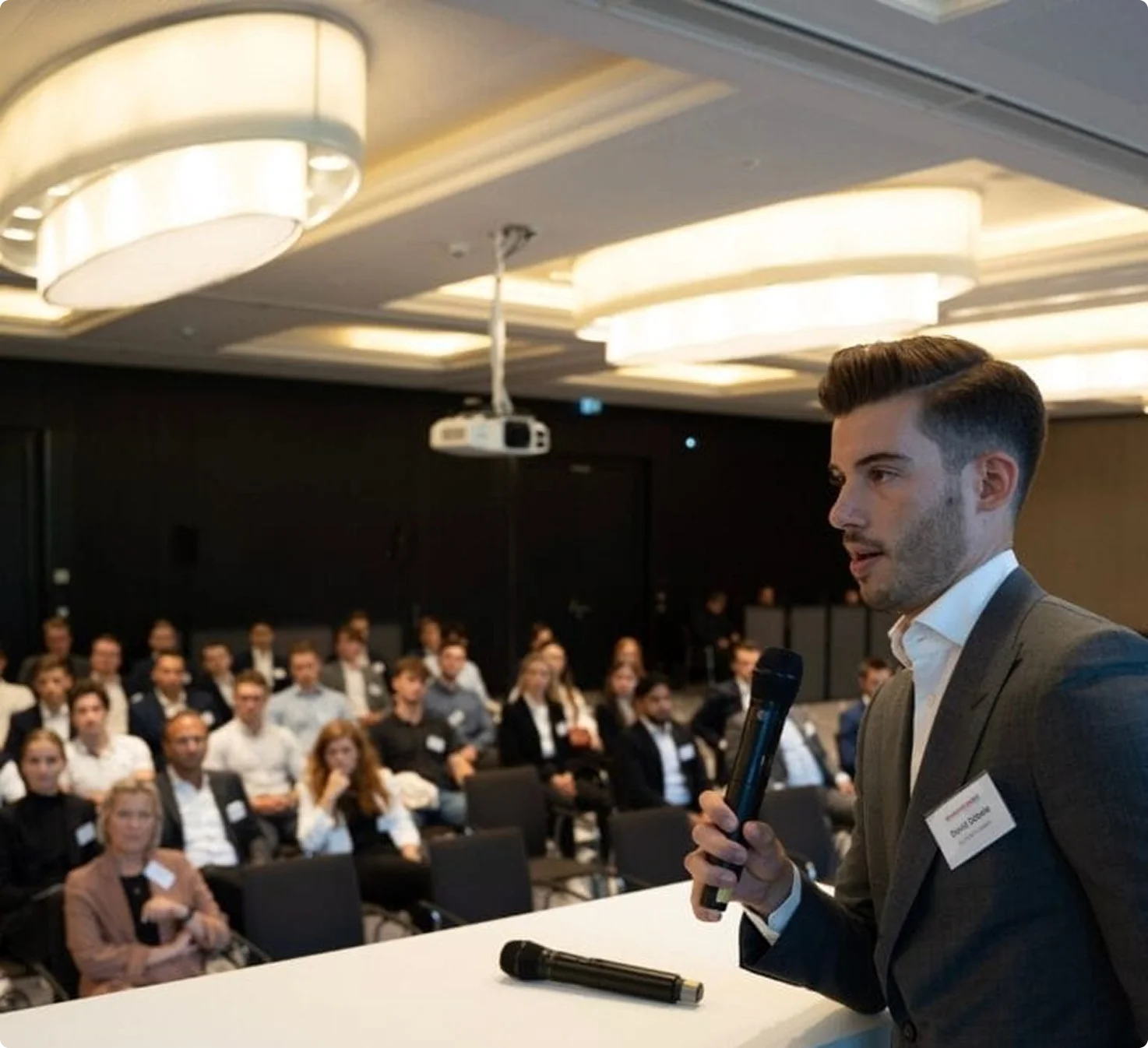 Junger Mann hält Mikrofon und spricht vor Publikum in einem Konferenzraum.