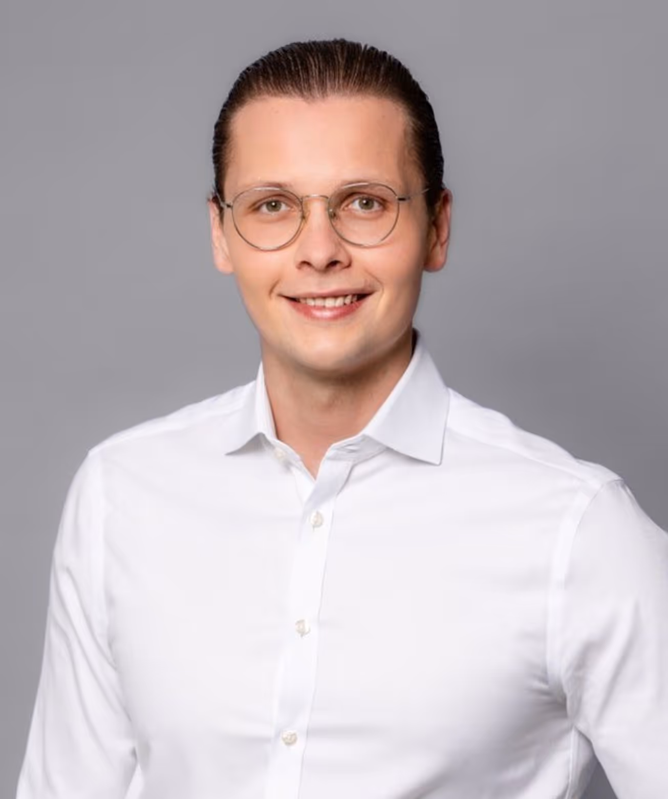 Lächelnder Mann mit Brille und zurückgebundenem Haar in weißem Hemd vor grauem Hintergrund.