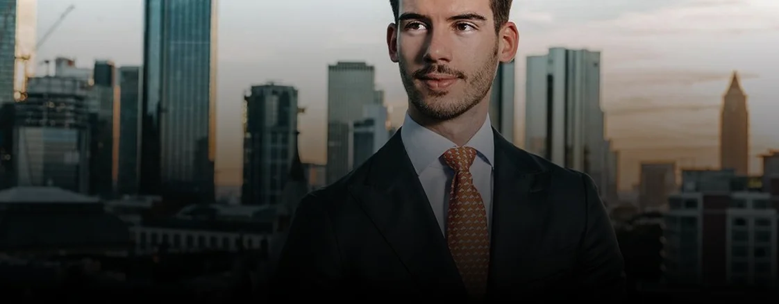 Junger Mann im Anzug mit orangefarbener Krawatte vor Hochhaus-Skyline bei Sonnenuntergang.