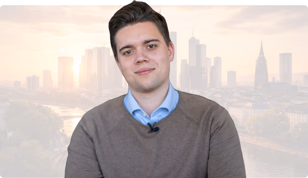Junger Mann mit braunem Pullover und blauem Hemd vor der Skyline einer Stadt bei Sonnenaufgang.