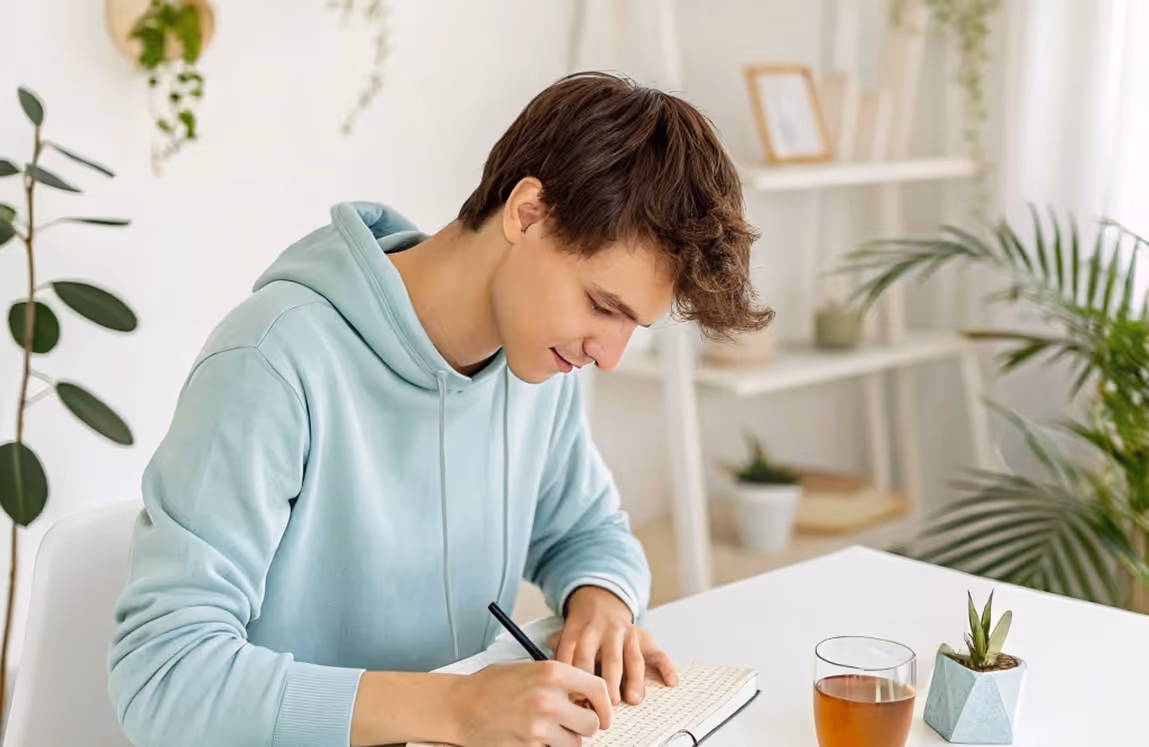 Junger Mann in blauem Hoodie schreibt in ein Notizbuch am Tisch mit Pflanze und Glas Tee.