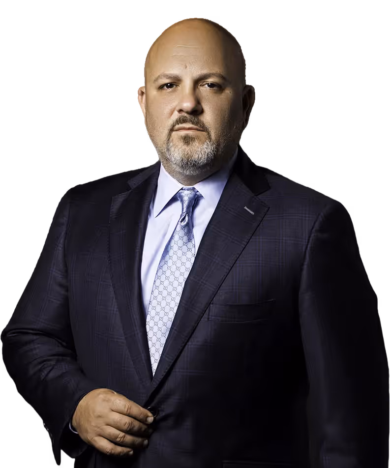 Serious bald man with a trimmed beard wearing a dark suit, light blue shirt, and patterned tie against a black background.