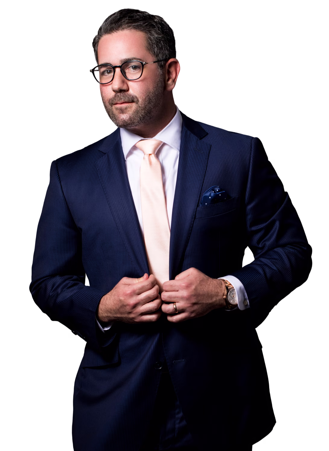 Man in a navy blue suit with a pale peach tie and glasses adjusting his jacket against a black background.