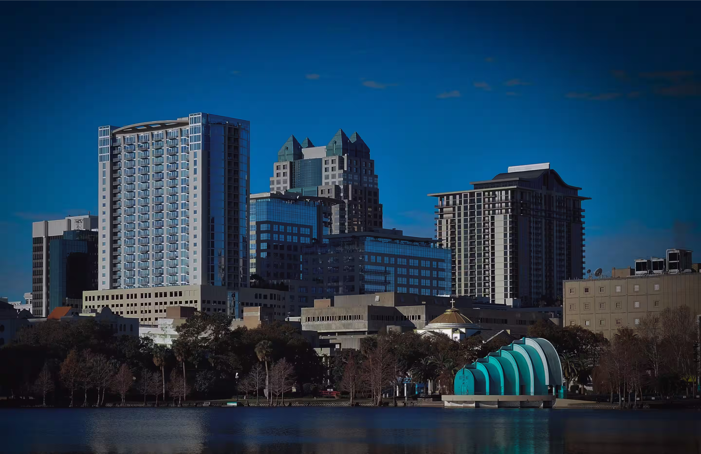 City skyline with modern high-rise buildings and a waterfront featuring a blue, shell-shaped structure under a clear blue sky.