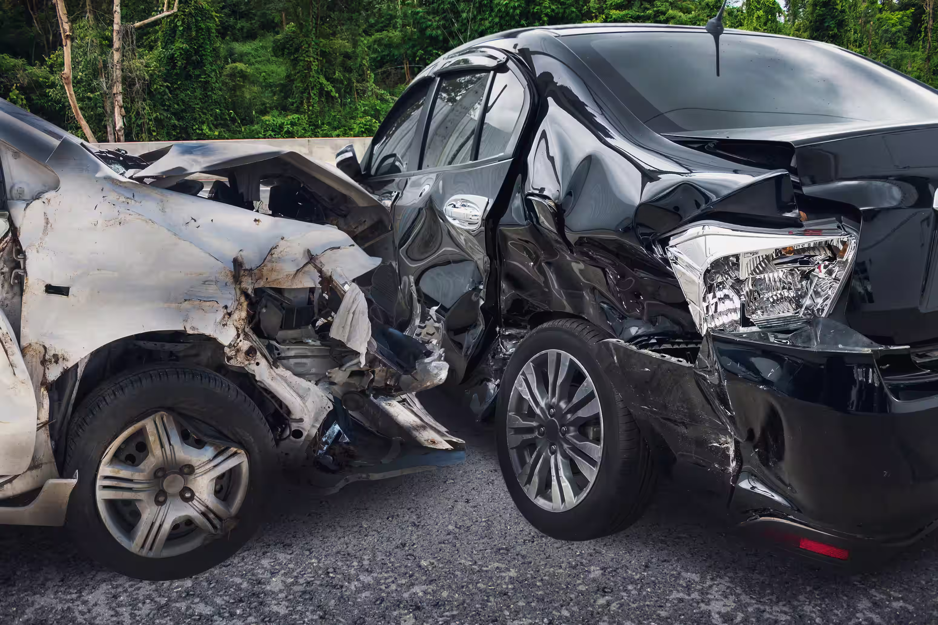 White and black cars with severe damage in a rear-end collision on a road with trees in the background.