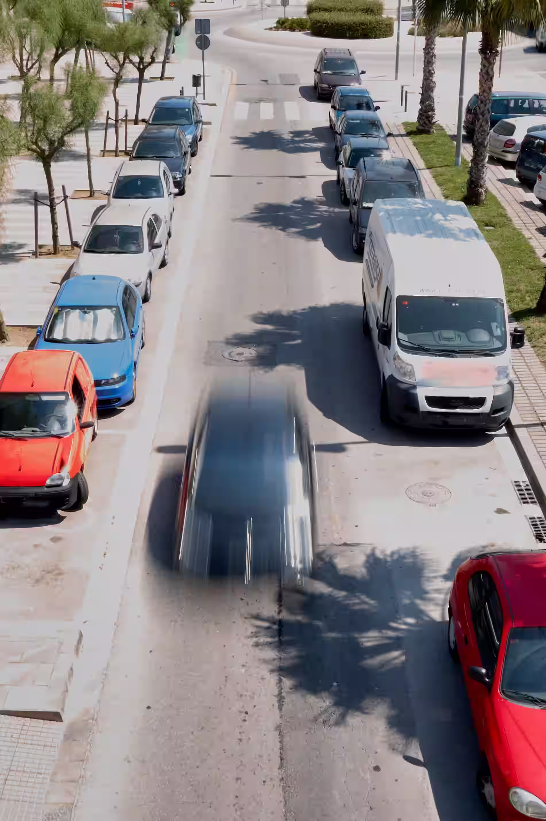 Street scene with parked cars on both sides and a blurred vehicle speeding down the road in bright daylight.