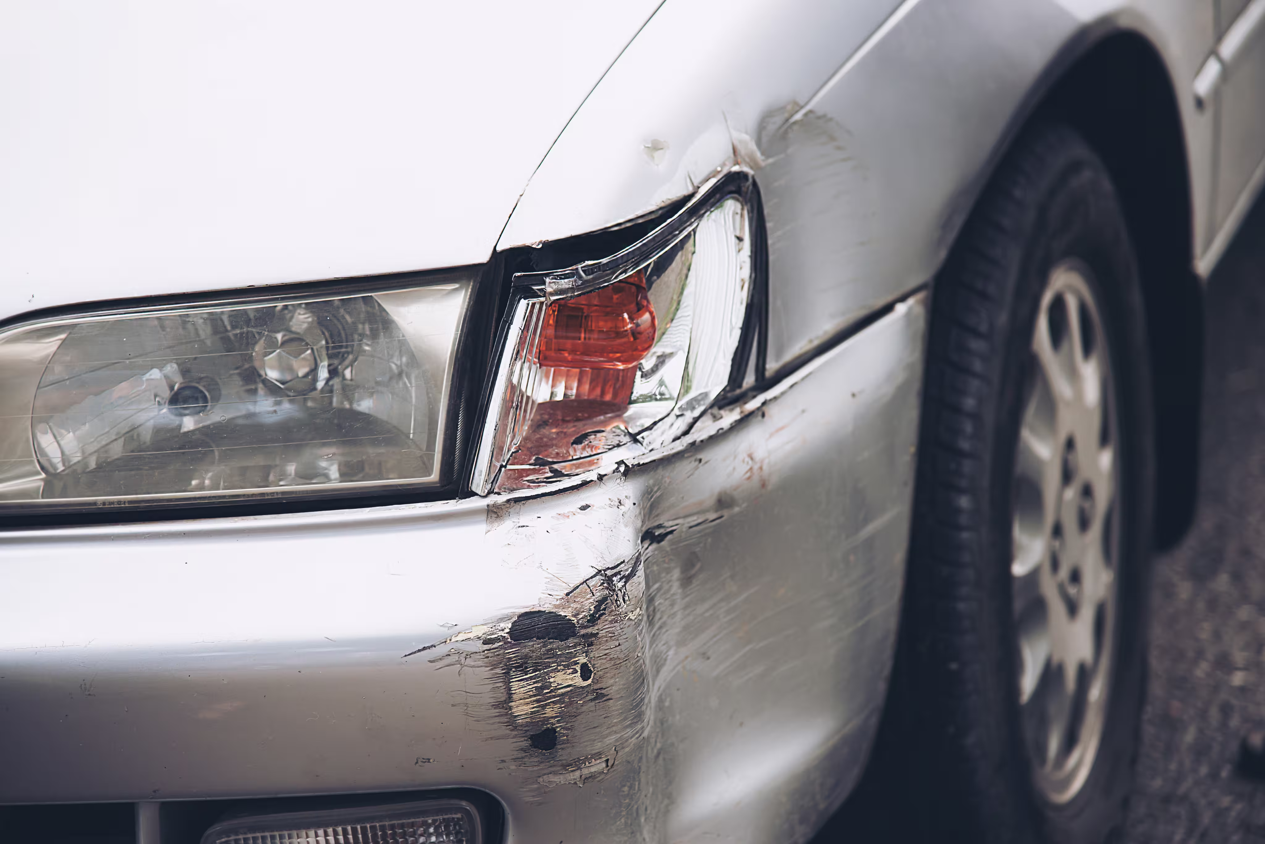 Close-up of a silver car's damaged front right side showing a cracked headlight and scrape marks on the bumper.