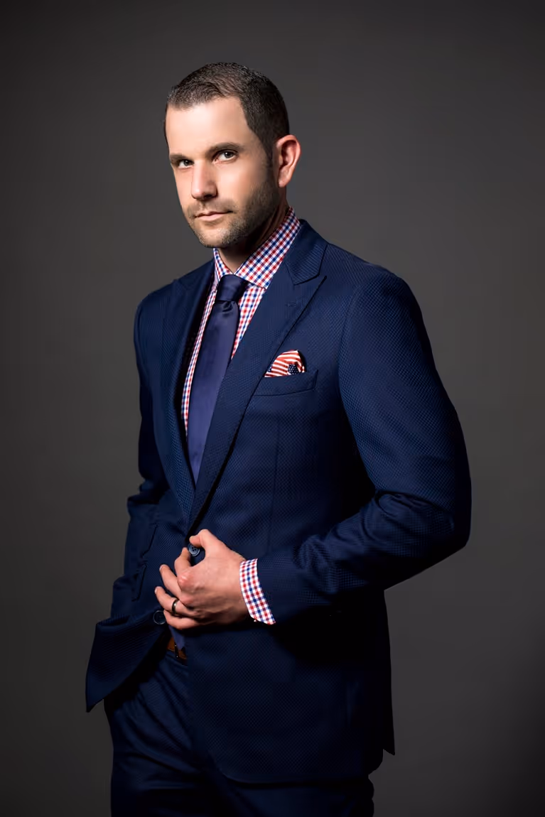 Man wearing a navy blue suit, patterned red and white shirt, and navy tie, posing against a dark gray background.