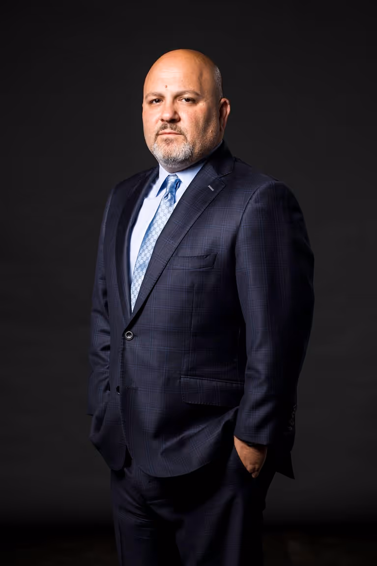 Bald man with a trimmed beard wearing a dark checkered suit and light blue patterned tie against a black background.