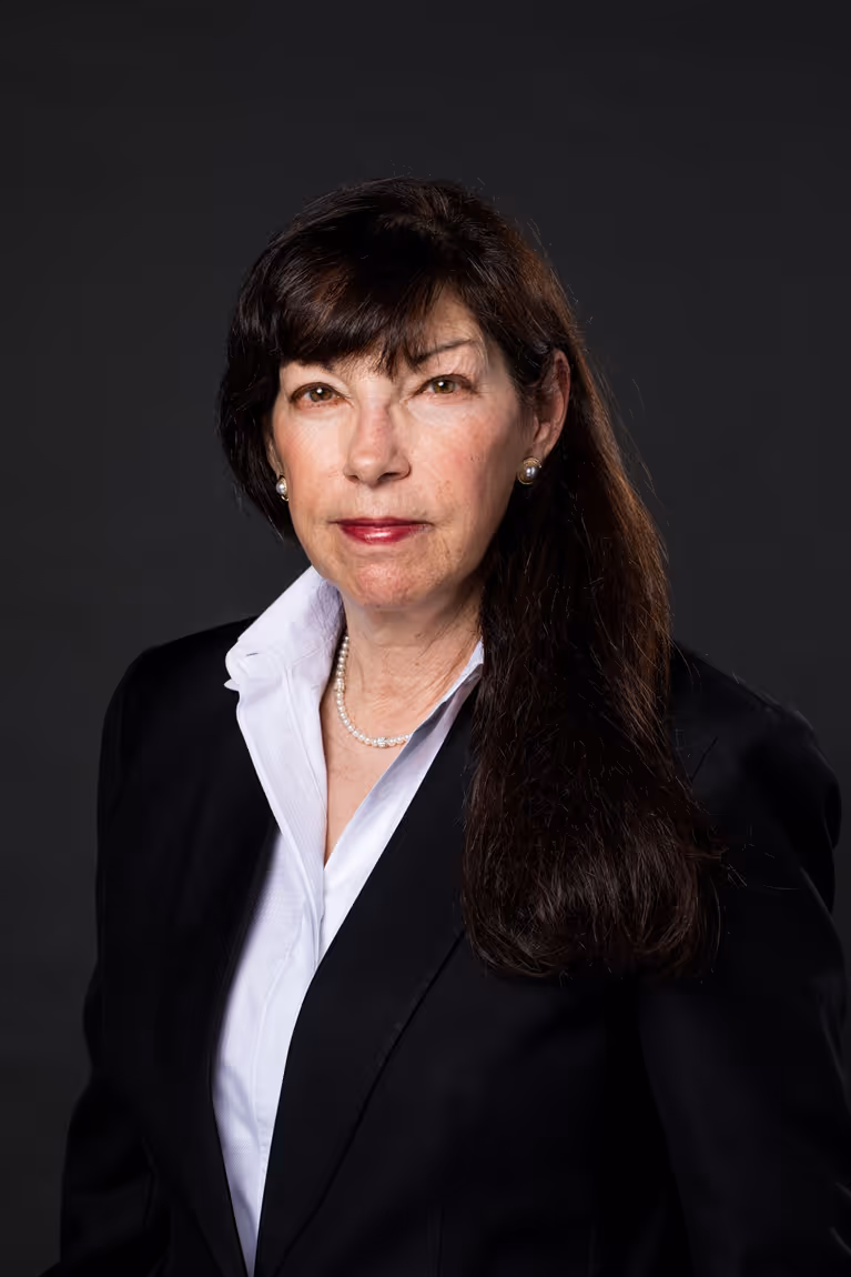 Professional woman with long dark hair wearing a black blazer, white shirt, pearl necklace, and pearl earrings against a dark background.