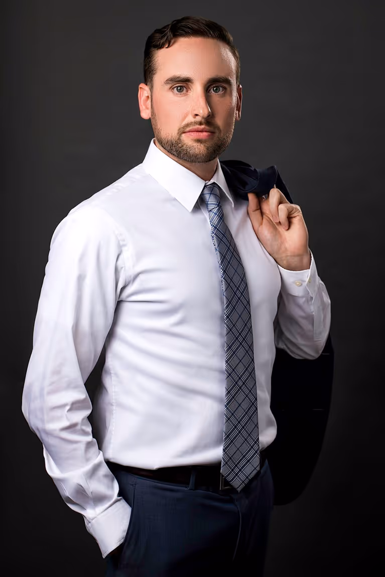Professional man in white dress shirt and plaid tie holding a dark jacket over his shoulder against a dark background.