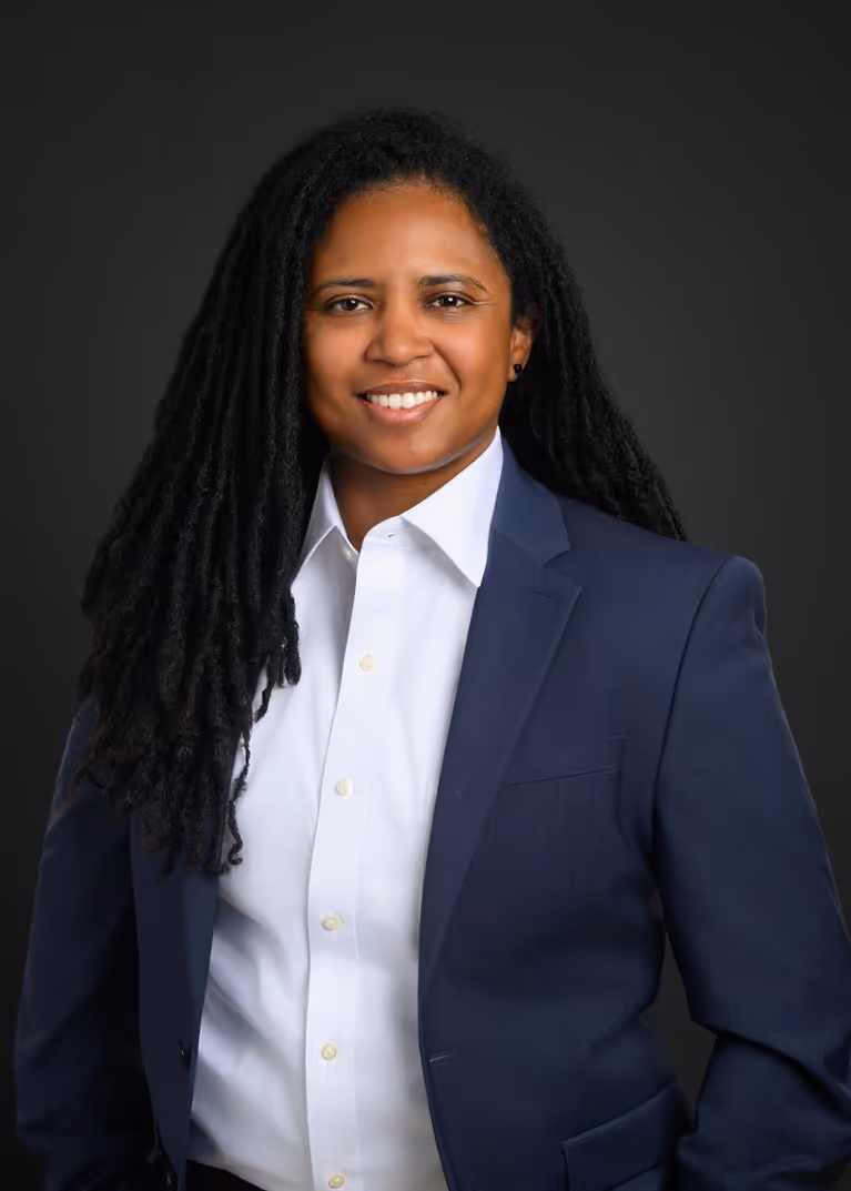 Smiling person with long dreadlocks wearing a white shirt and navy blazer against a dark background.