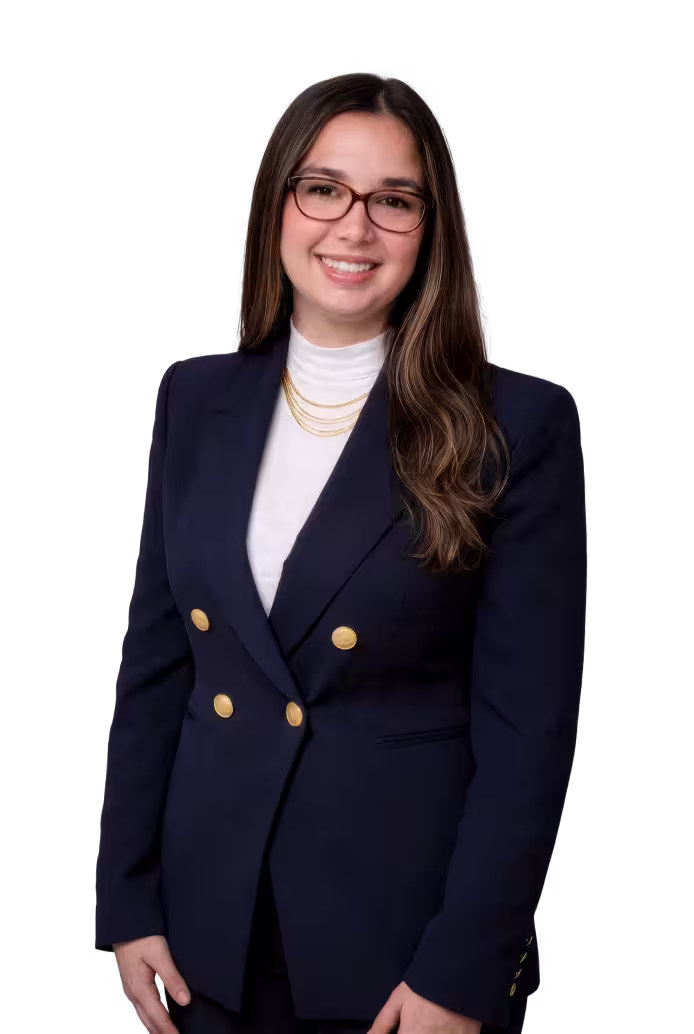 Smiling woman with long brown hair, wearing glasses, a navy blazer with gold buttons, a white high-neck top, and layered gold necklaces.