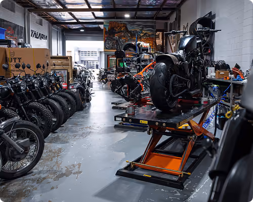 Motorcycle workshop with bikes lined on the left and a bike on a lift in the center under a transparent roof.