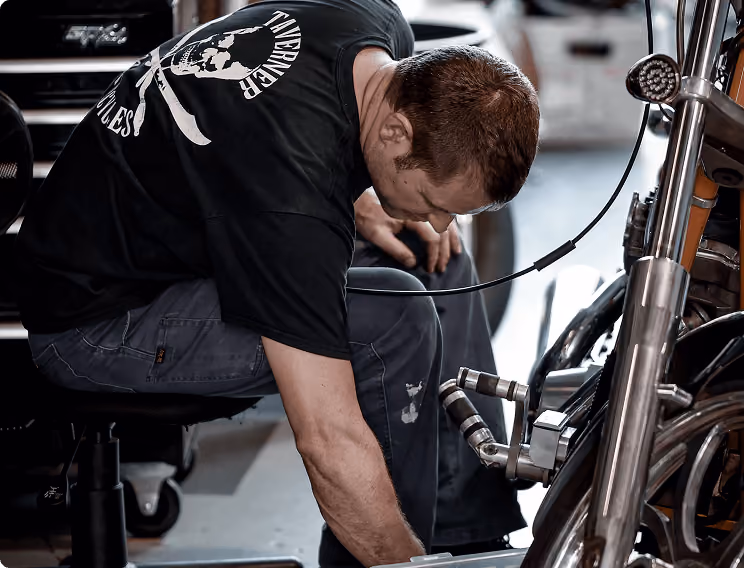 Man in black t-shirt working on a motorcycle in a garage or workshop.