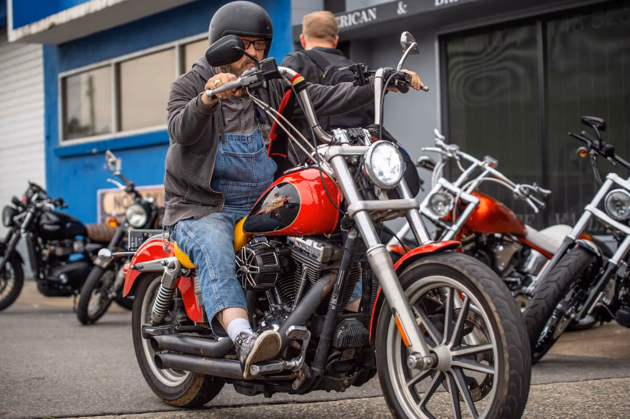 Man wearing a helmet rides a red Harley-Davidson motorcycle in front of a building with other bikes parked.