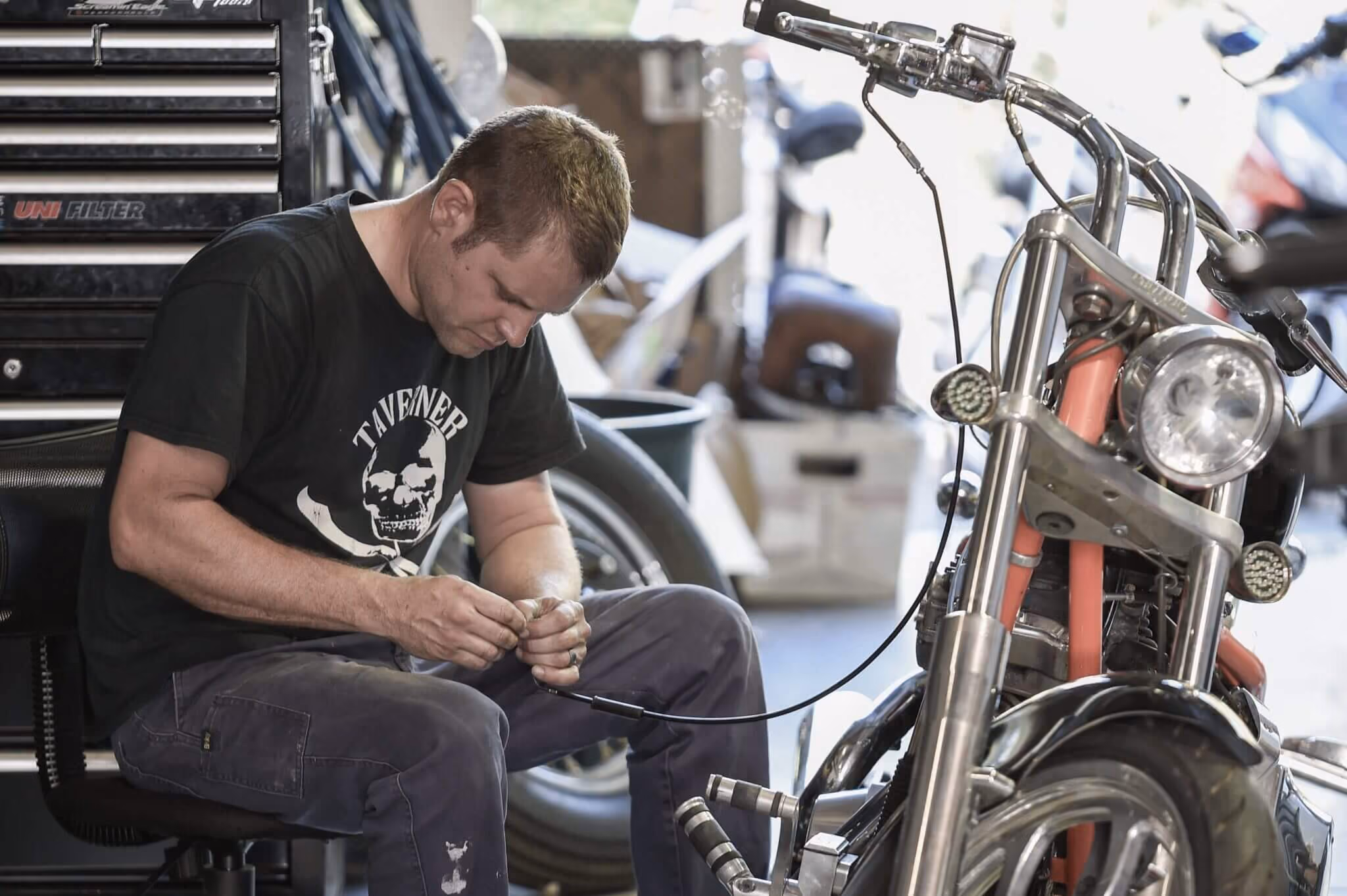 Man sitting in a workshop, repairing a cable near a motorcycle front wheel and handlebars.