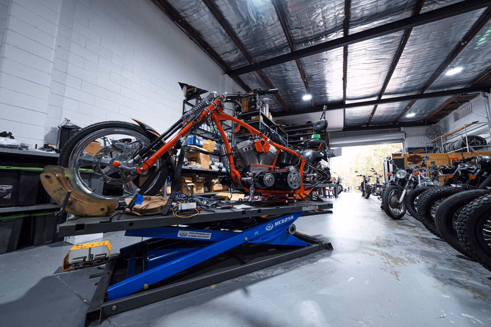 Custom motorcycle on a blue hydraulic lift in a workshop with tires and bikes in the background.