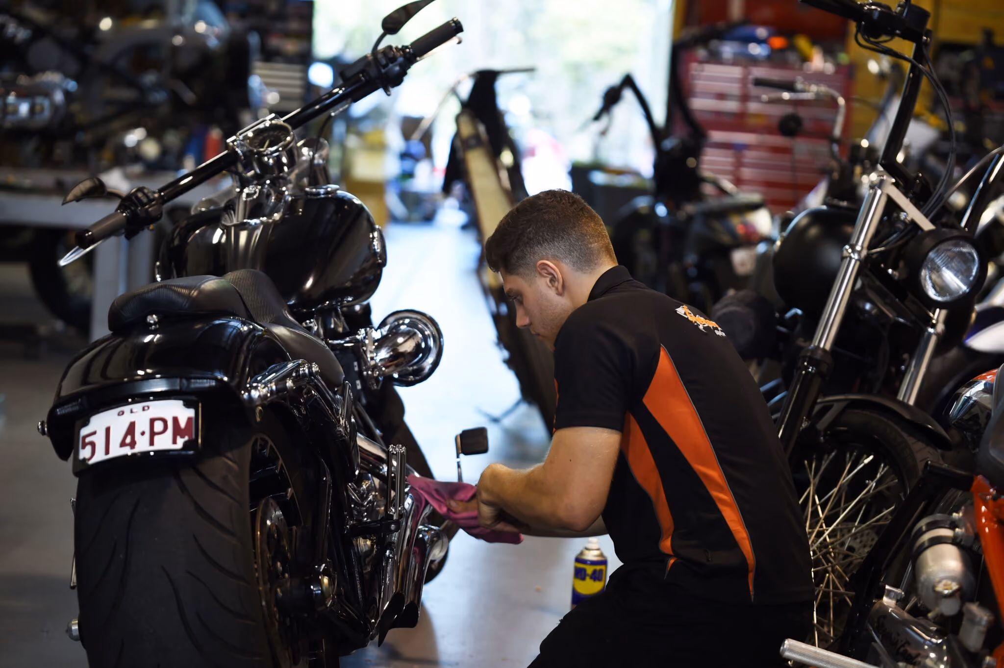 Person cleaning or repairing a black motorcycle in a workshop with tools and other bikes nearby.
