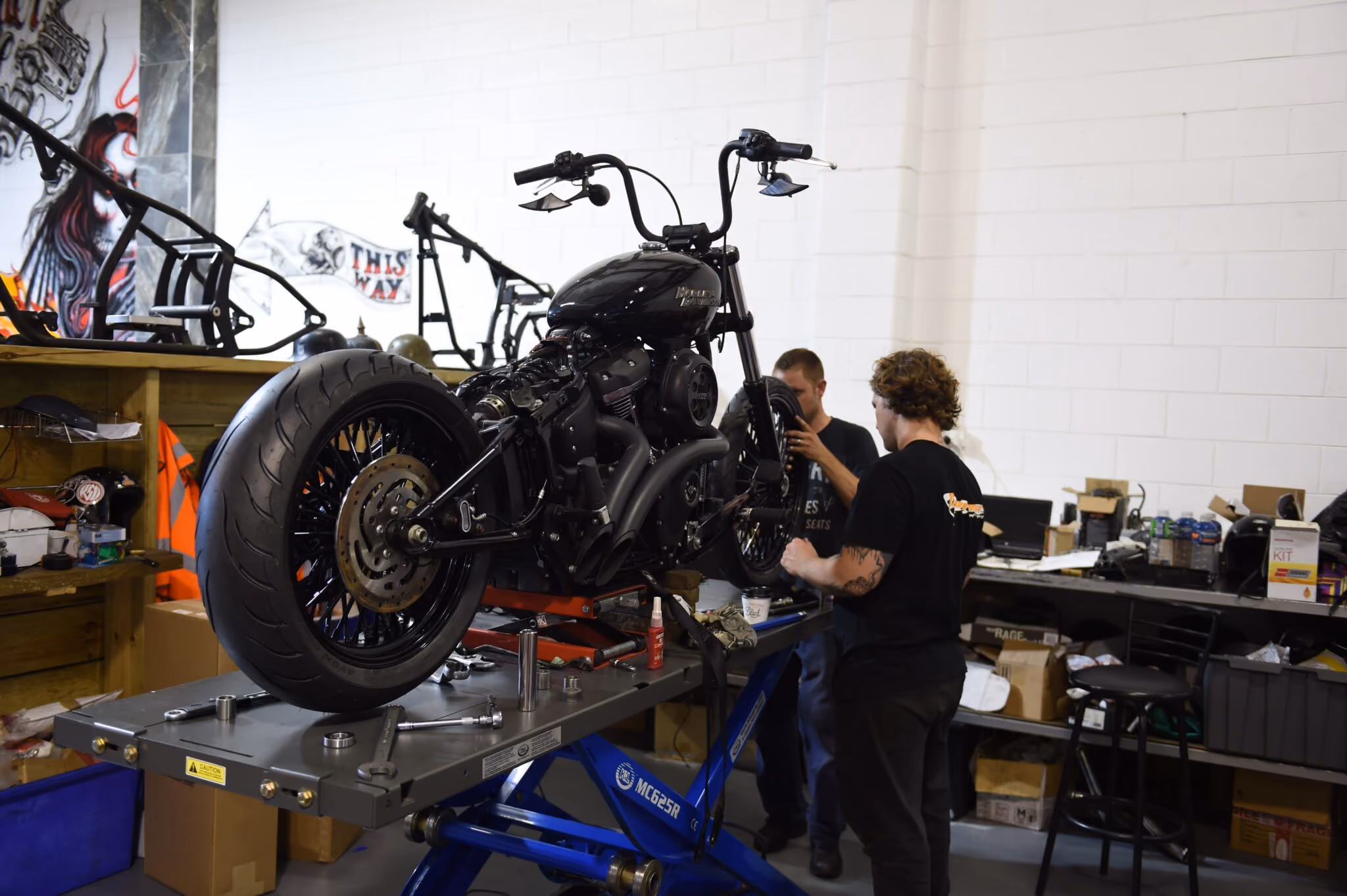 Two mechanics working on a black motorcycle elevated on a blue hydraulic lift in a garage.