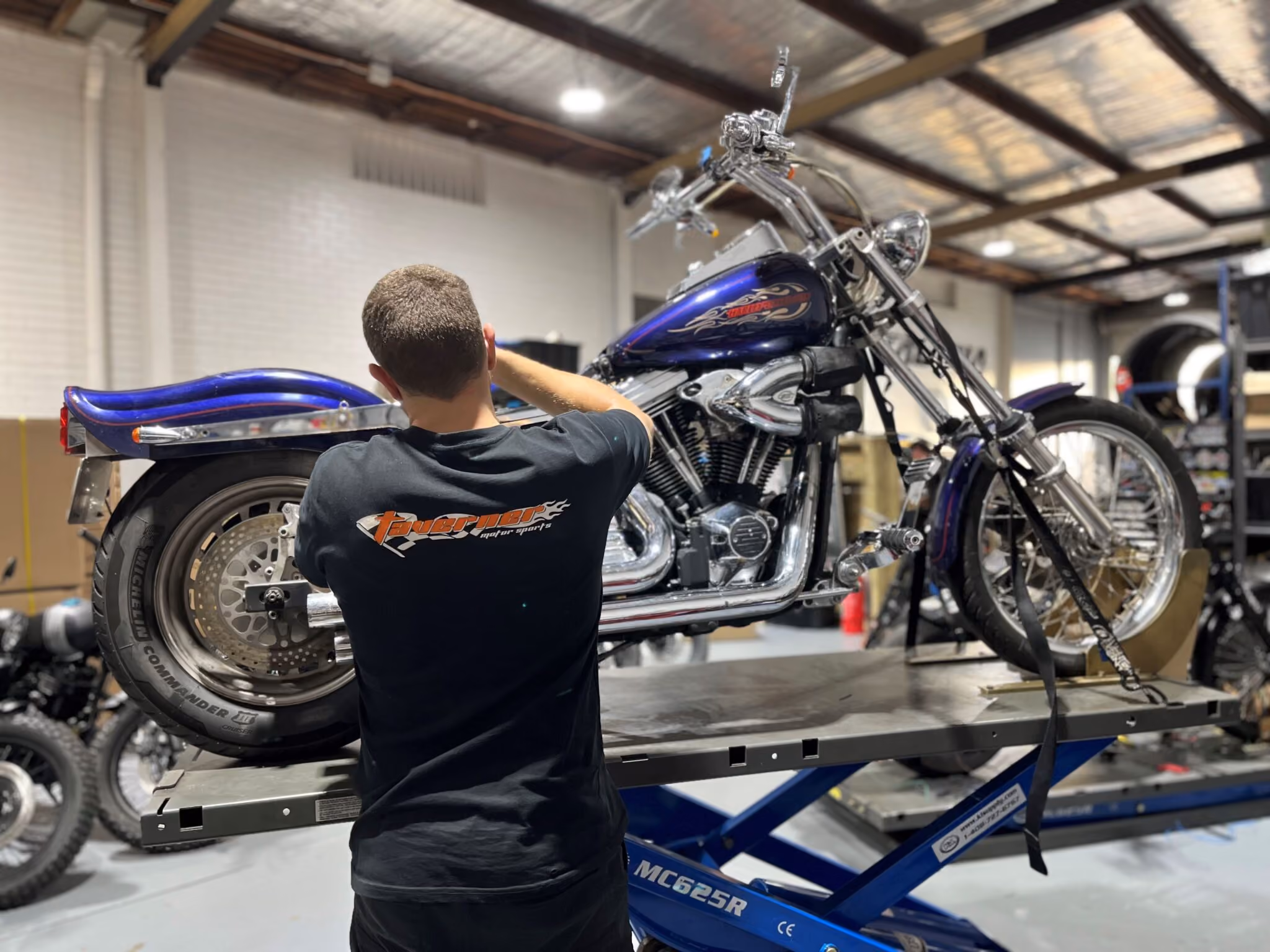 Technician working on a purple motorcycle mounted on a blue repair lift in a workshop.