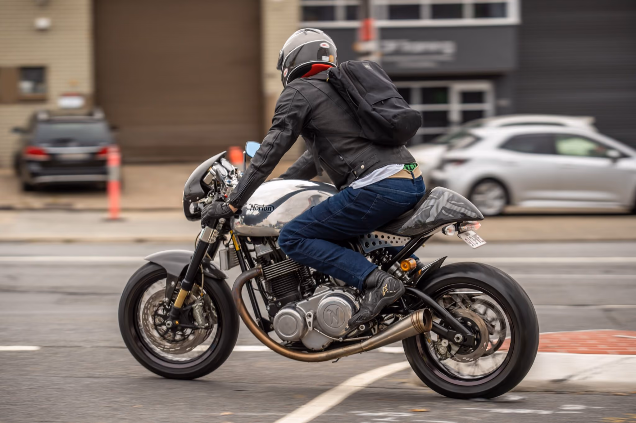 Person wearing a helmet and leather jacket riding a silver Norton motorcycle on a city street.