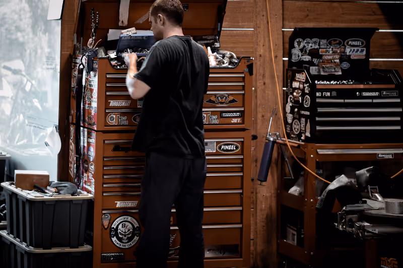 Man in black shirt working in a workshop with tool cabinets covered in stickers.