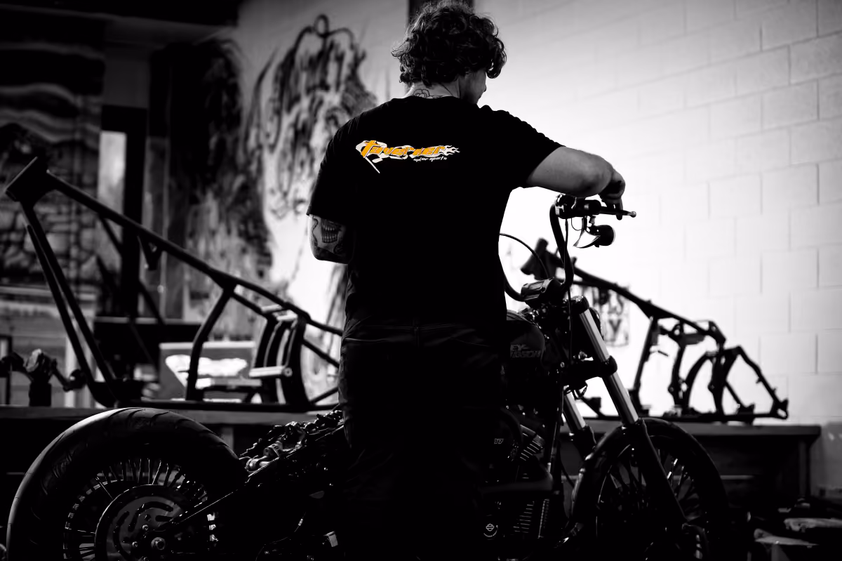 Man with curly hair working on a motorcycle in a workshop with bike frames in the background.