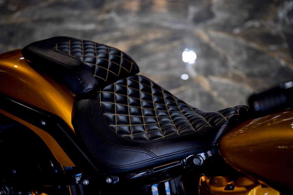 Black leather motorcycle seat with yellow stitching on a metallic orange motorcycle body.