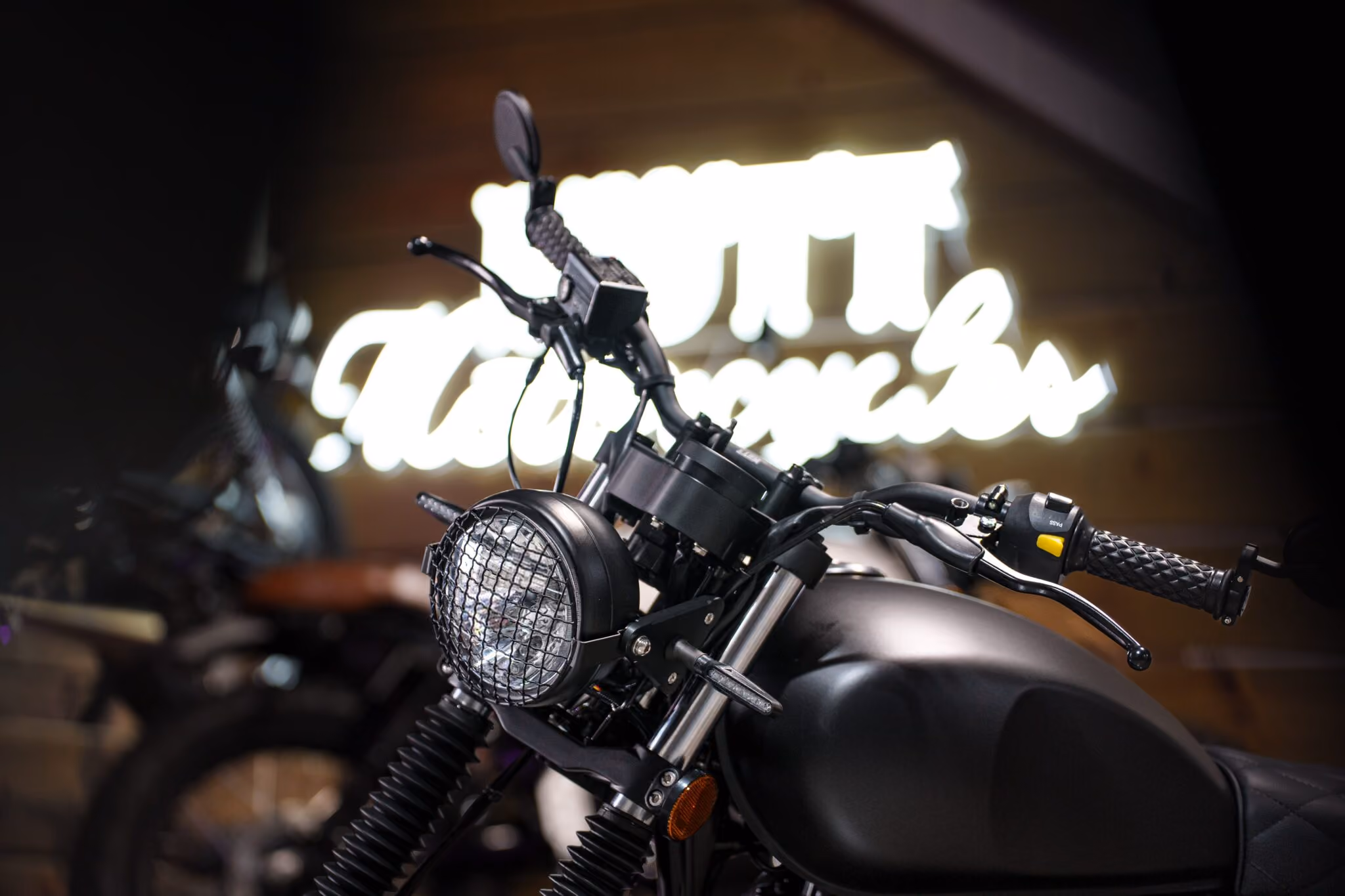 Close-up of a black motorcycle's front headlight, handlebars, and gas tank with a blurred neon sign in the background.