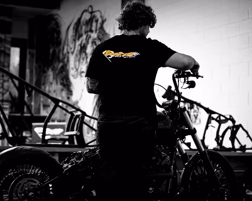 Man in black shirt standing next to a motorcycle in a dim indoor space with graffiti on the wall.