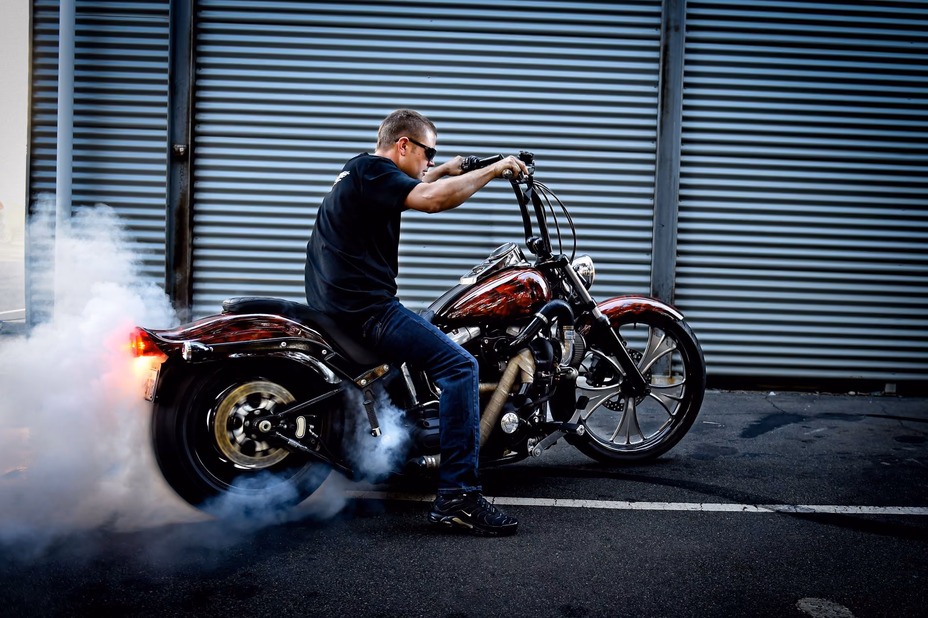 Person in sunglasses doing a burnout on a custom motorcycle near a corrugated metal wall.