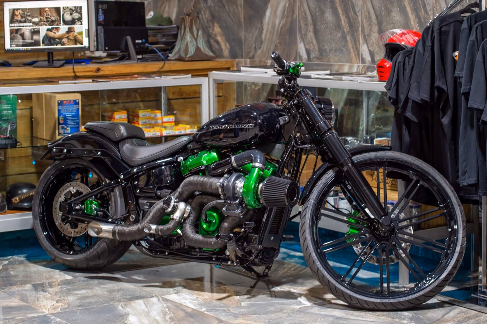 Black custom Harley-Davidson motorcycle with green engine parts displayed indoors near racks and a monitor.