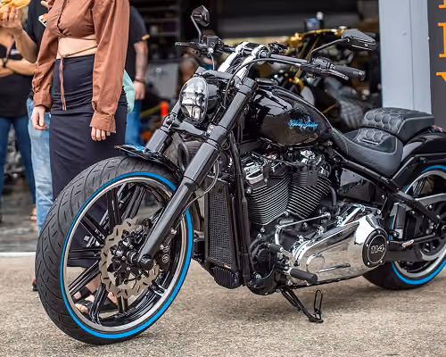 Black custom motorcycle with blue trim on wheels parked on pavement, people standing in background.